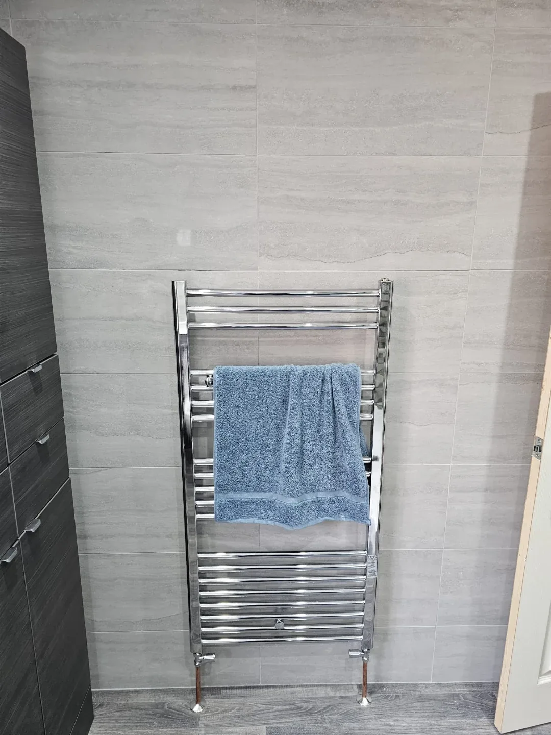 Chrome heated towel rail fitted against newly tiled walls in a completed bathroom.