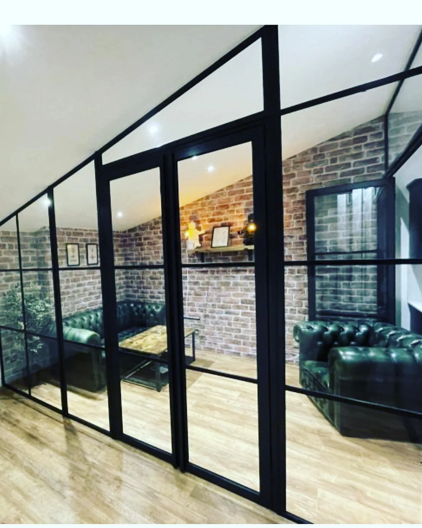 Internal black steel-framed glazed screen and doors dividing a snug with an exposed brick wall and a green leather chesterfield sofa.