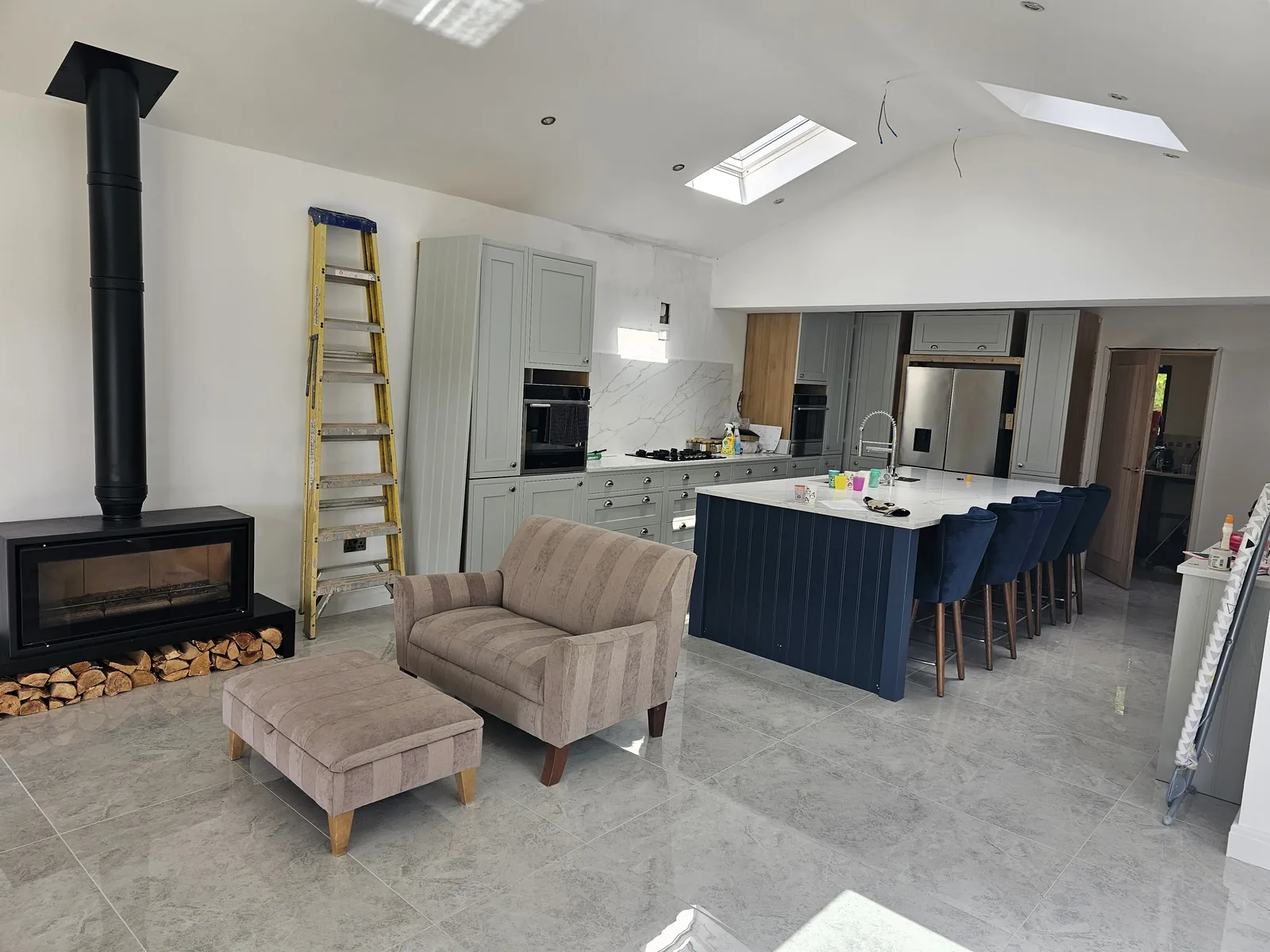 Vaulted open-plan kitchen and living space with a navy island and bar stools, a log burner and roof windows above.