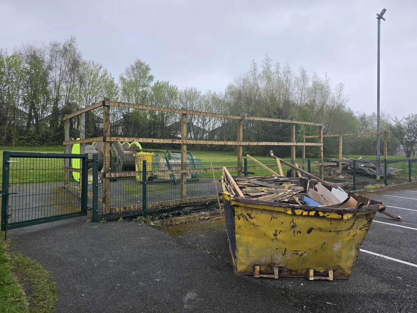 School grounds being cleared, with a skip of removed timber and old play-area framework behind green mesh fencing.