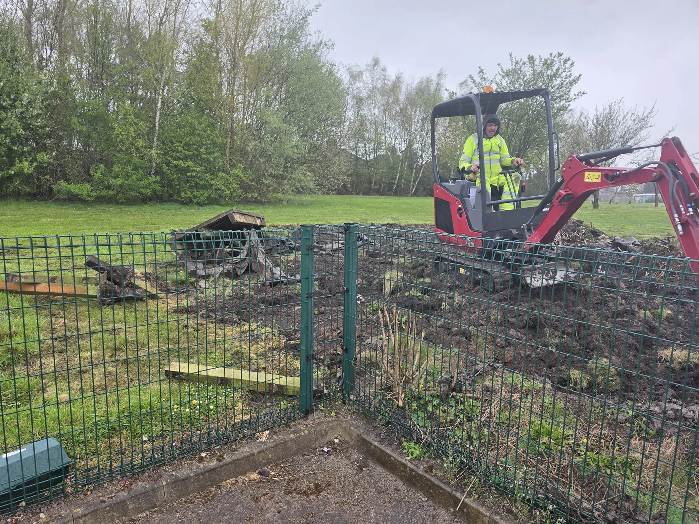Mini digger carrying out groundworks on the school field behind green mesh fencing.