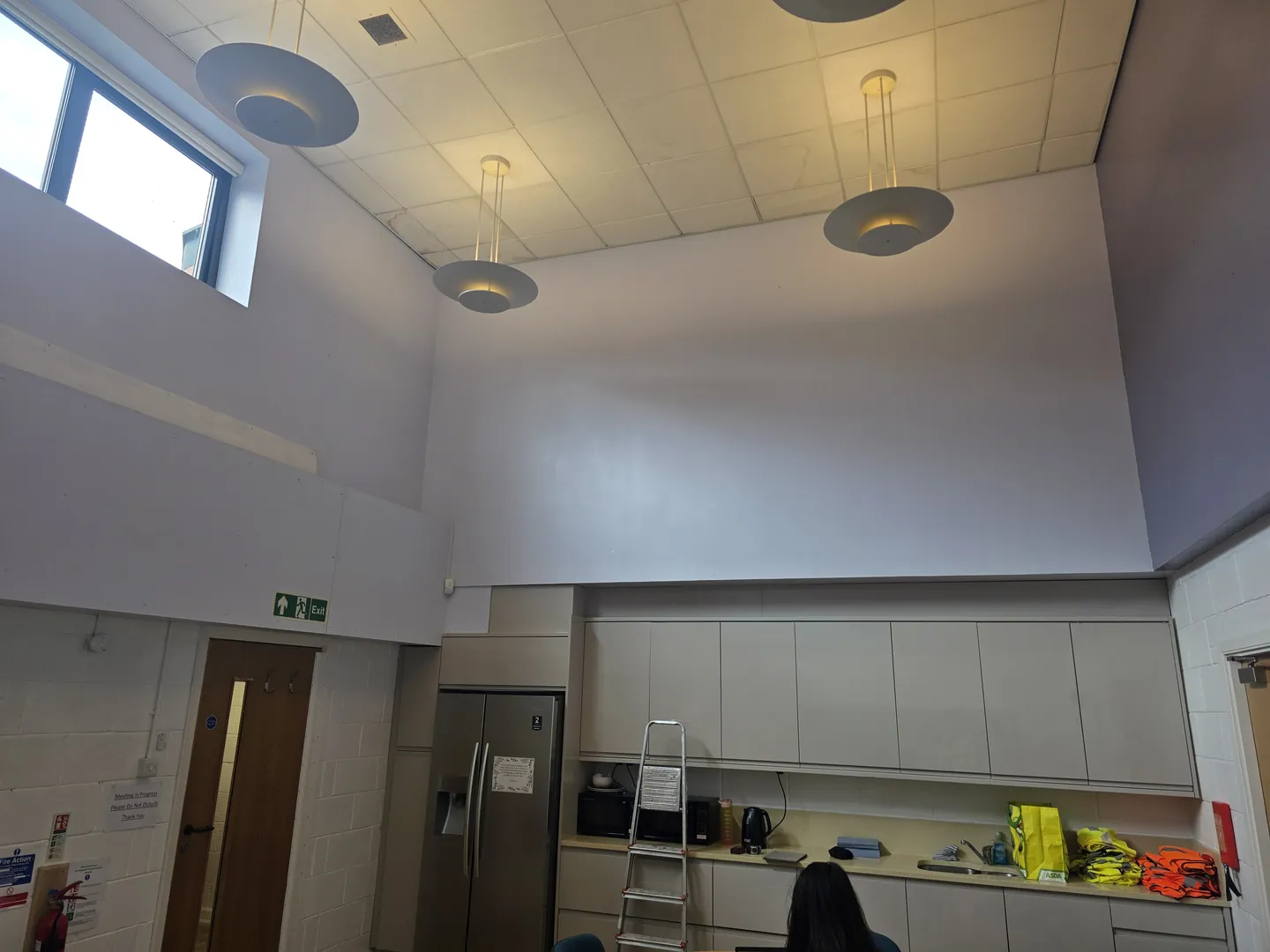 Nursery kitchen area under a high ceiling with new pendant lights, fitted base and wall units and a fridge, during final fit-out.