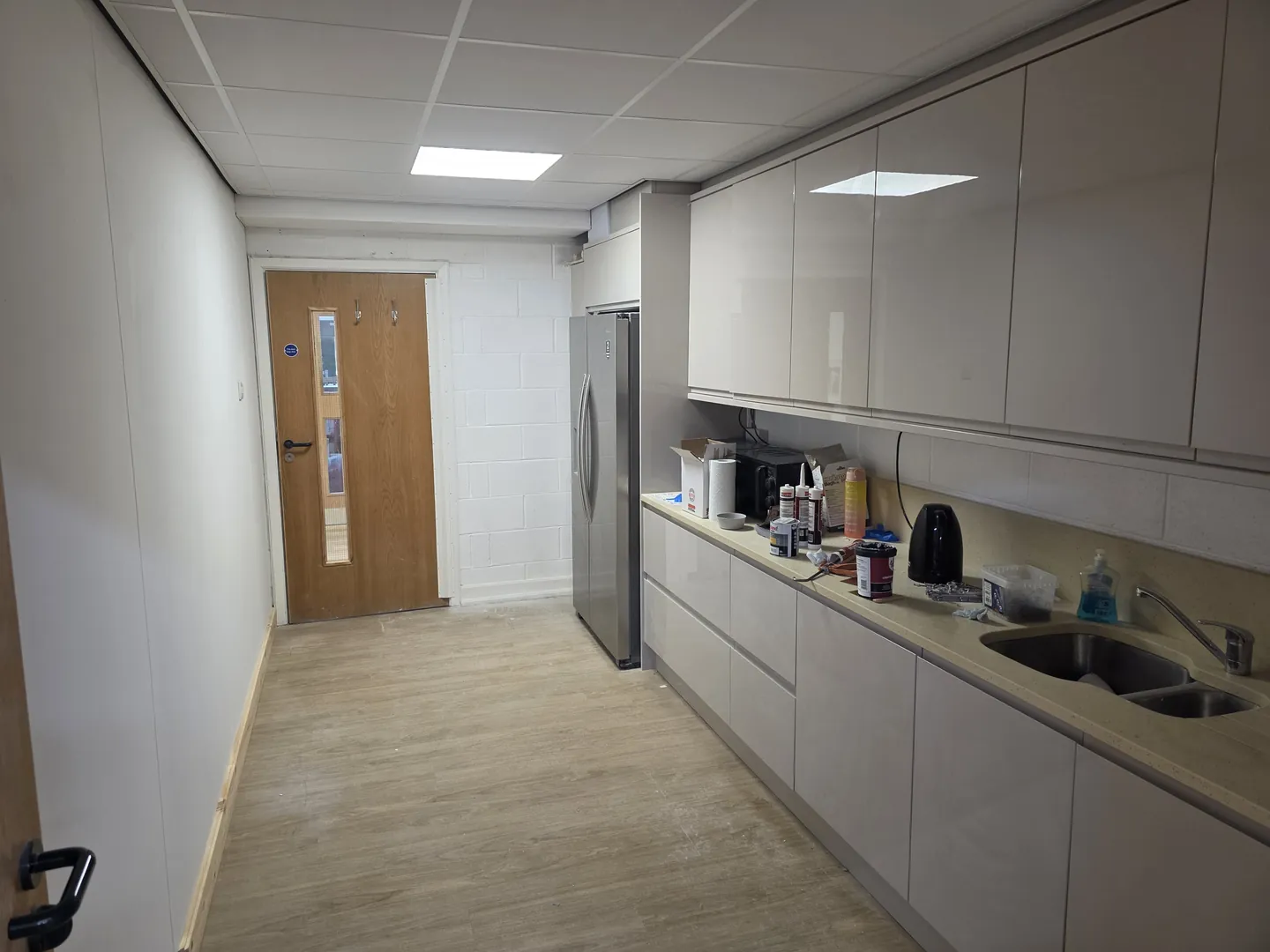 Completed school staff kitchen run along a new corridor, with gloss units, worktop, sink, LVT flooring and recessed ceiling lights.
