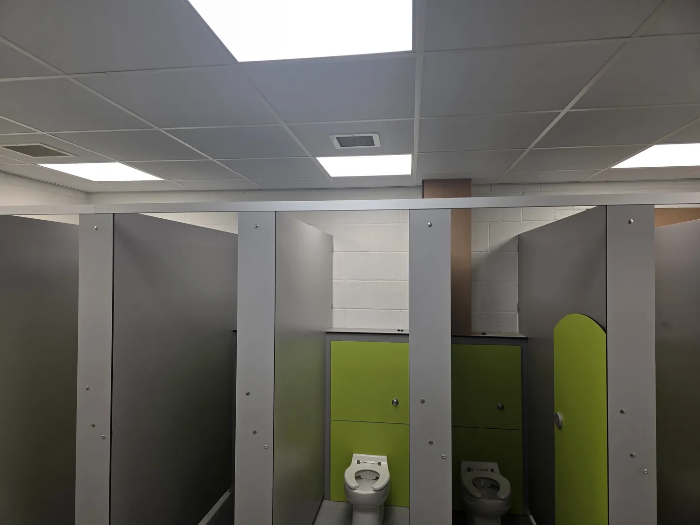 New nursery WC block with grey cubicle partitions, green doors, low-level toilets and a new suspended ceiling with LED panels.